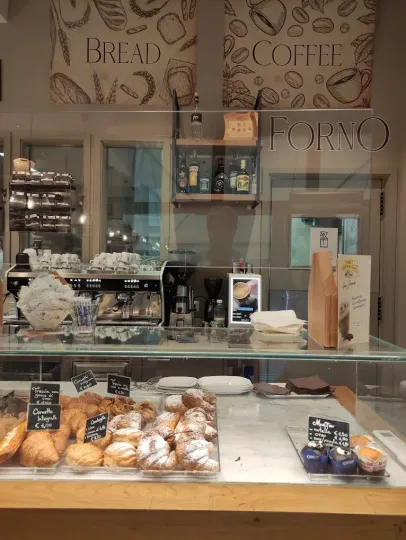 Dehor esterno del caffè con tavoli e sedie sotto un grande ombrellone, decorato con fiori rosa in vasi di legno.