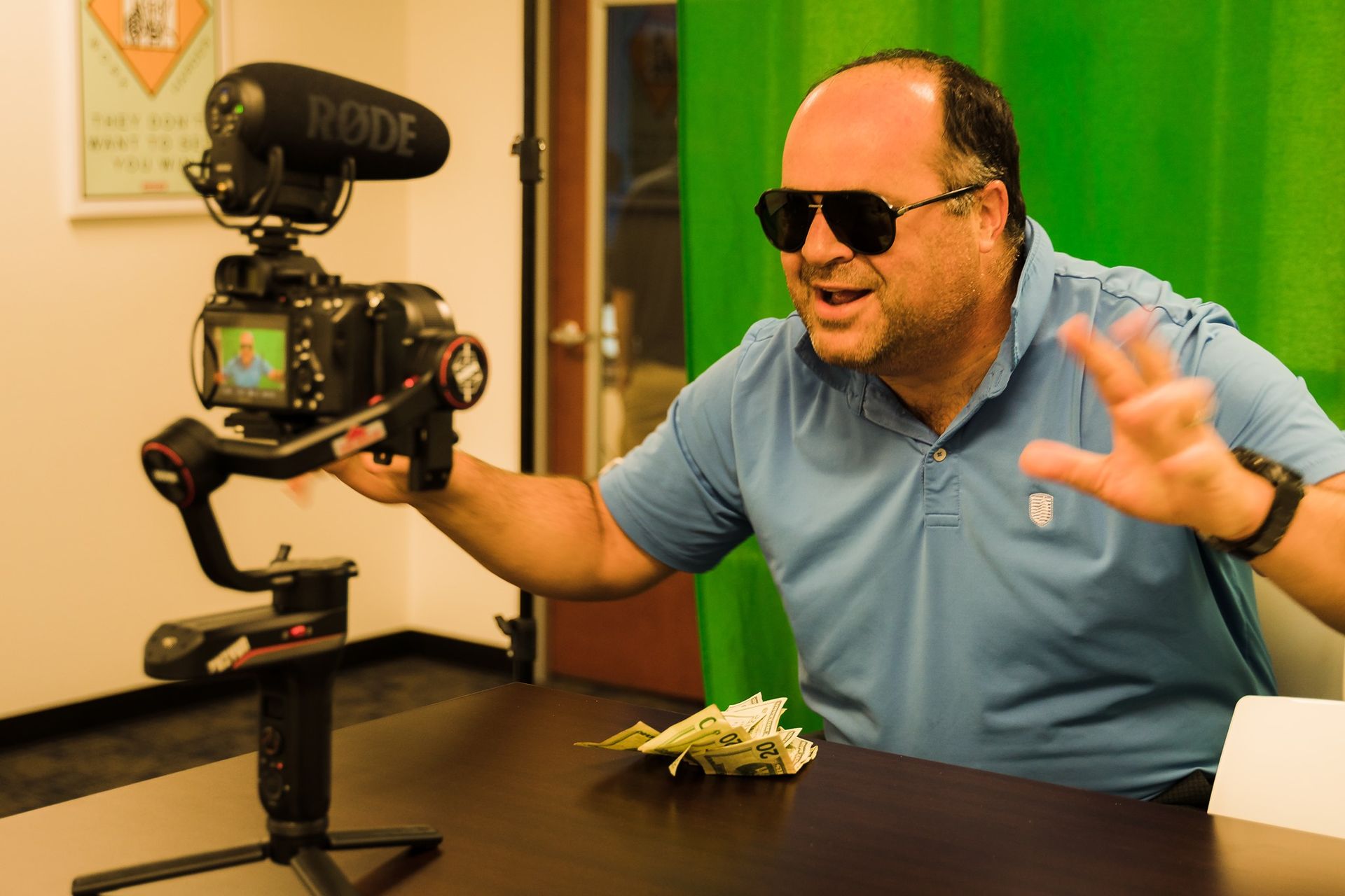 A man wearing sunglasses is holding a rode camera in front of a green screen.