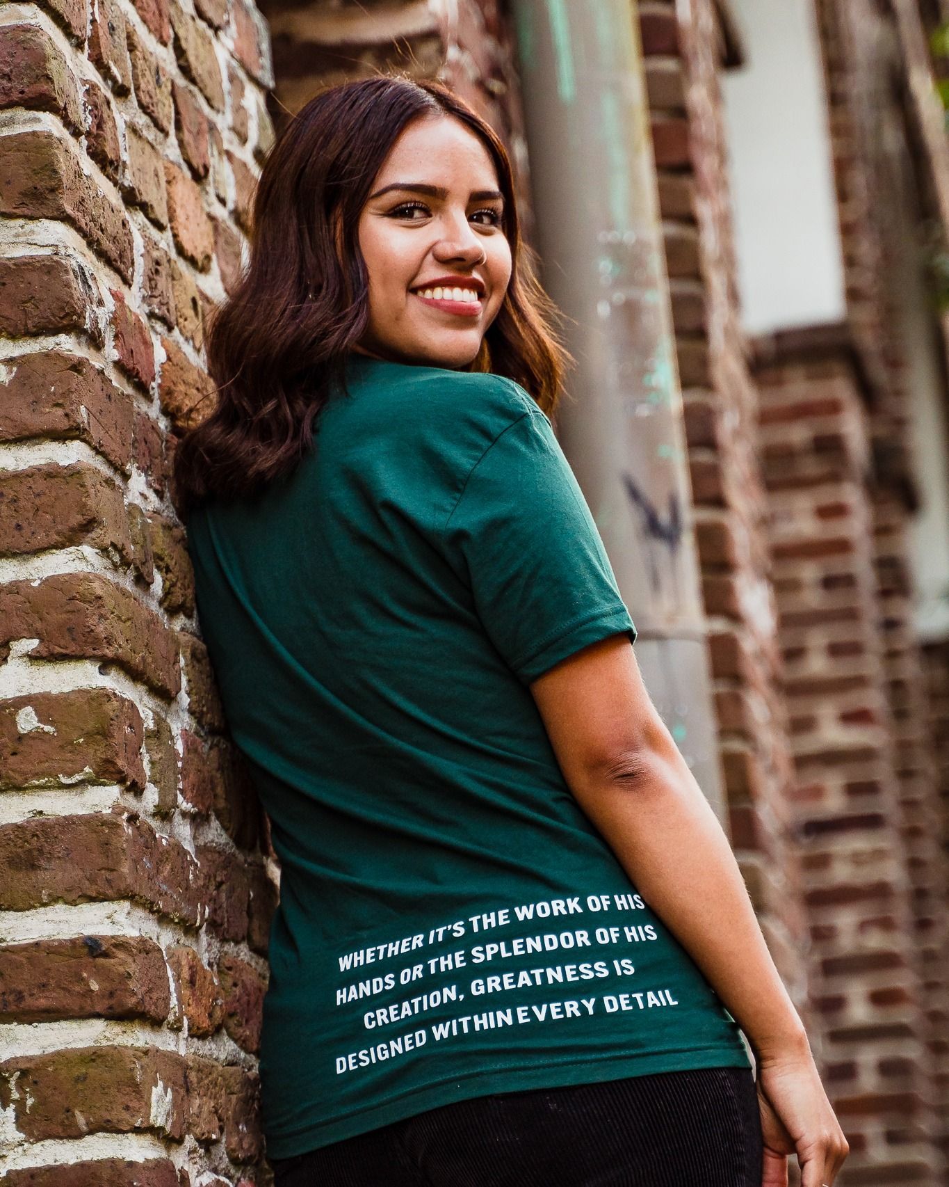A woman is leaning against a brick wall wearing a green t-shirt.