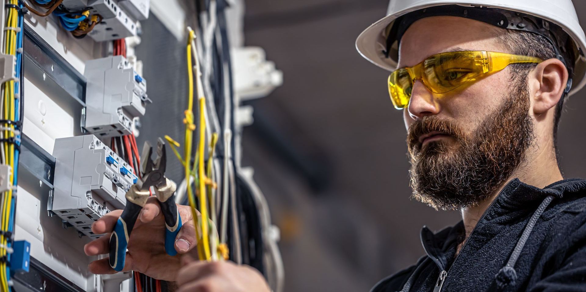 A man with a beard is wearing safety glasses and a hard hat while working on an electrical box.