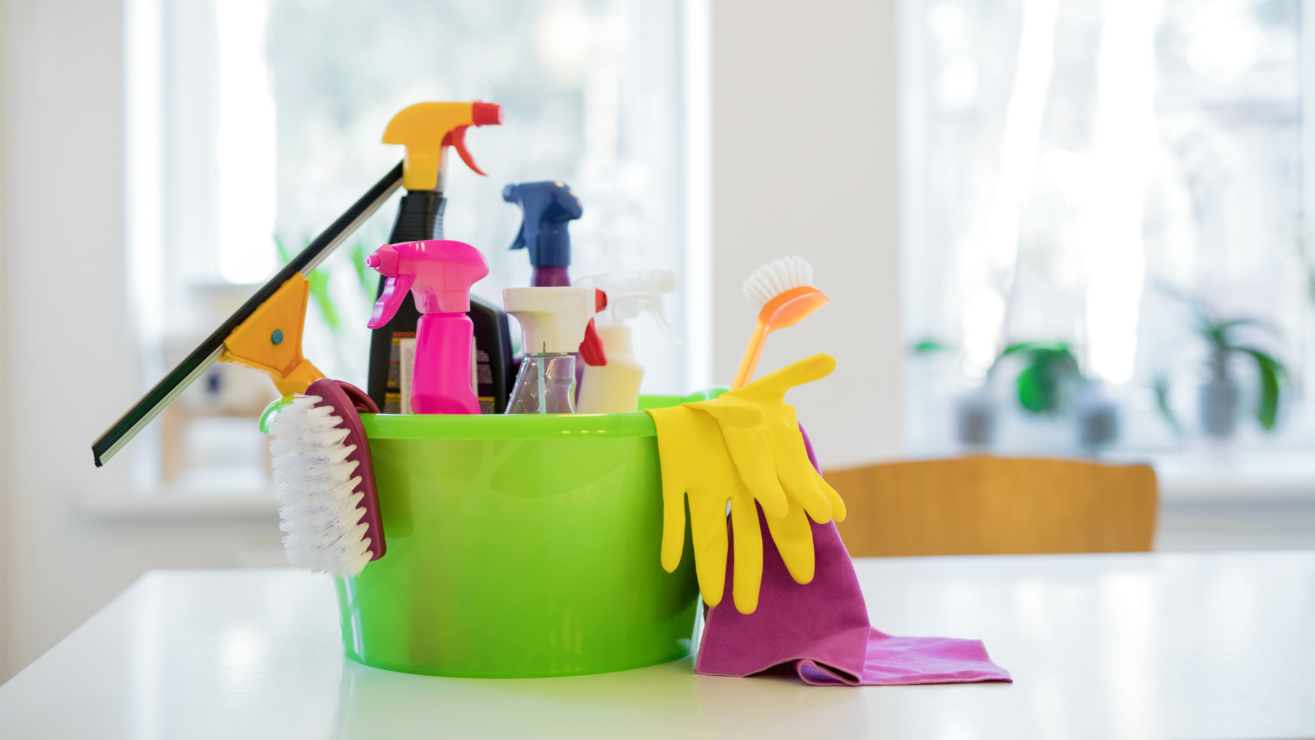 Green bucket of cleaning supplies on a white table: spray bottles, brushes, gloves, window cleaner.