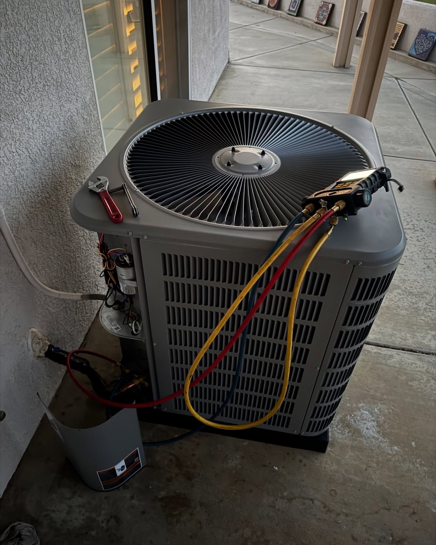 AC unit with gauges and hoses attached, tools nearby. Gray unit, yellow and red hoses, outdoors.