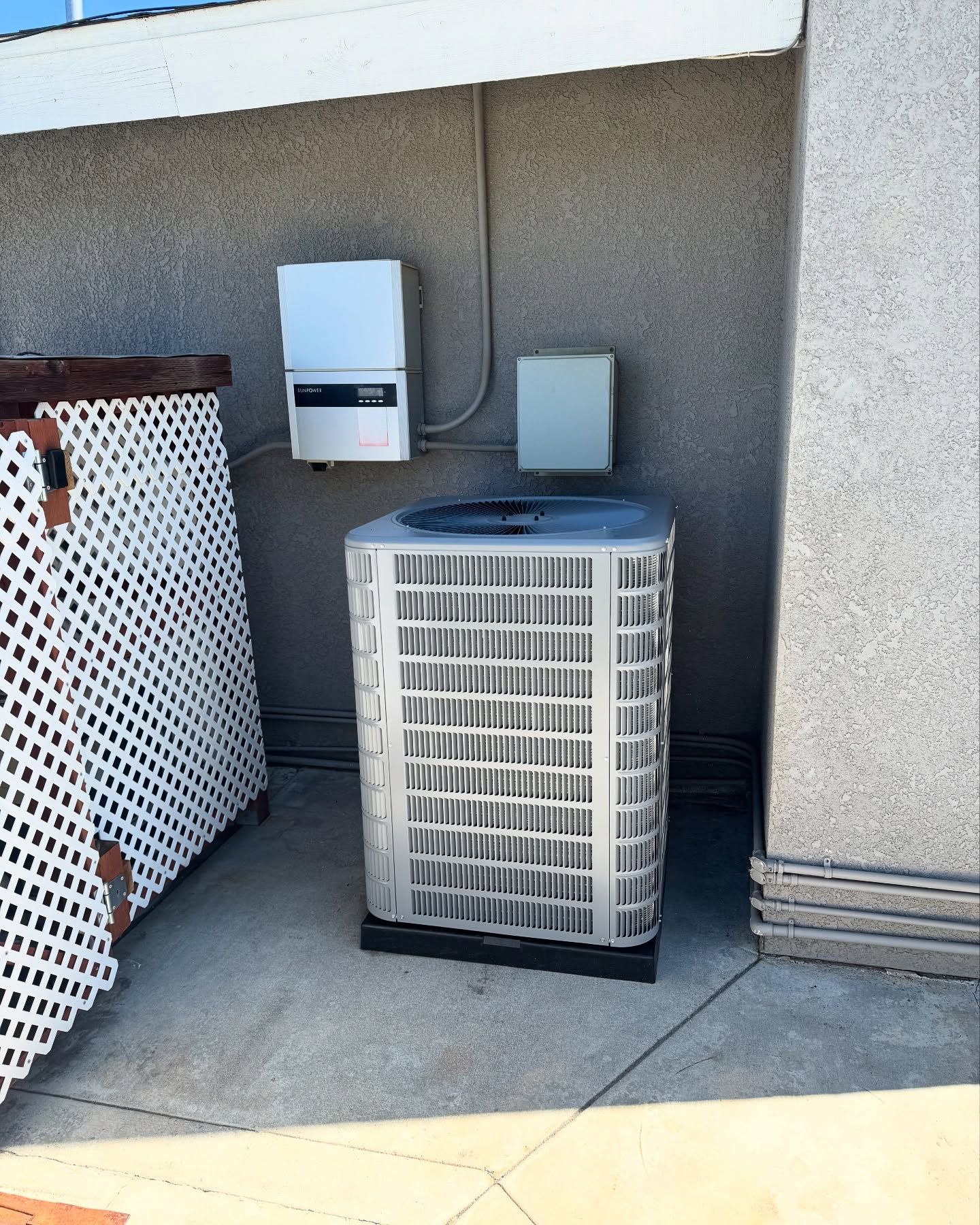 Air conditioning unit outdoors with electrical boxes, set against a stucco wall, near a lattice fence.
