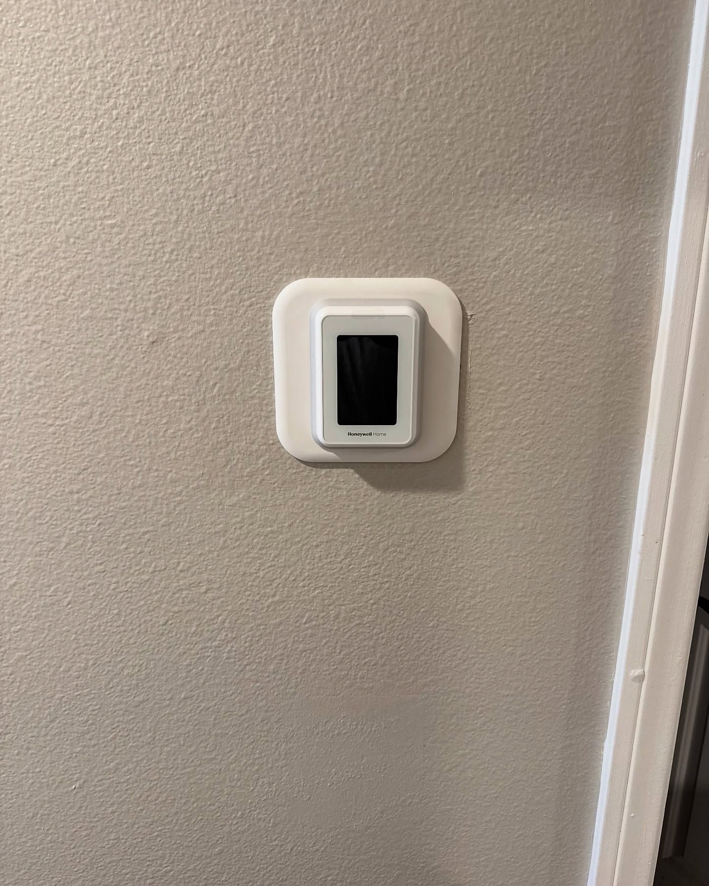 Thermostat with black screen on a textured off-white wall, next to a white door frame.