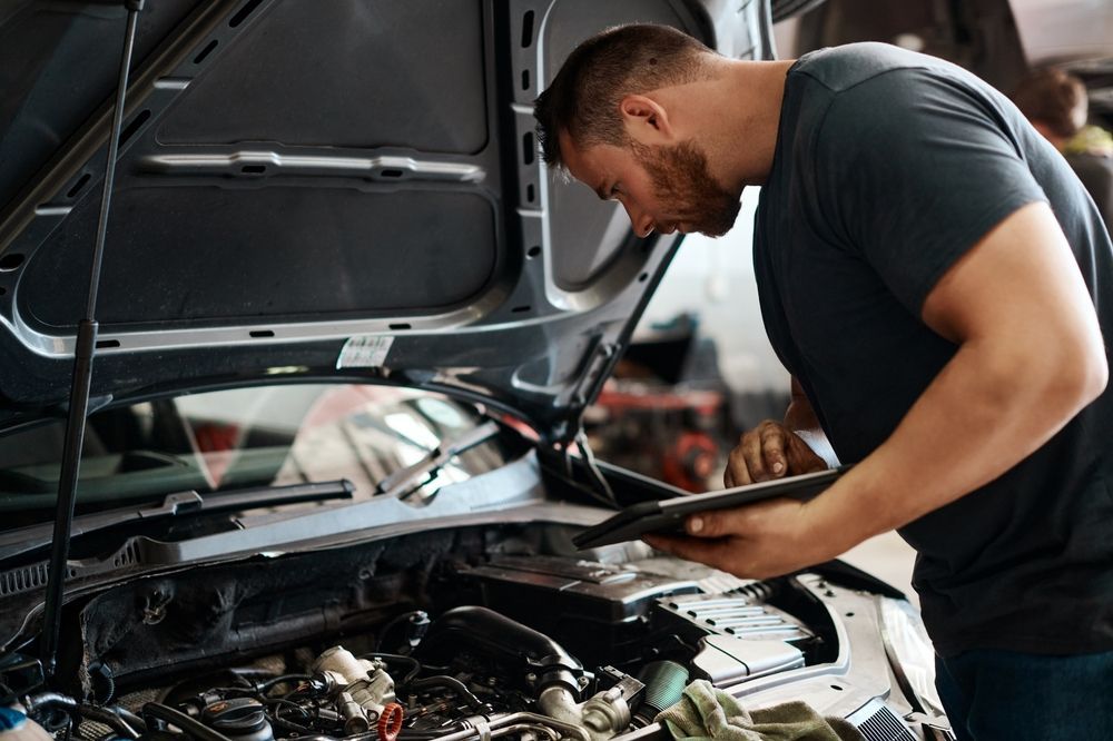 A man is looking under the hood of a car while holding a tablet.