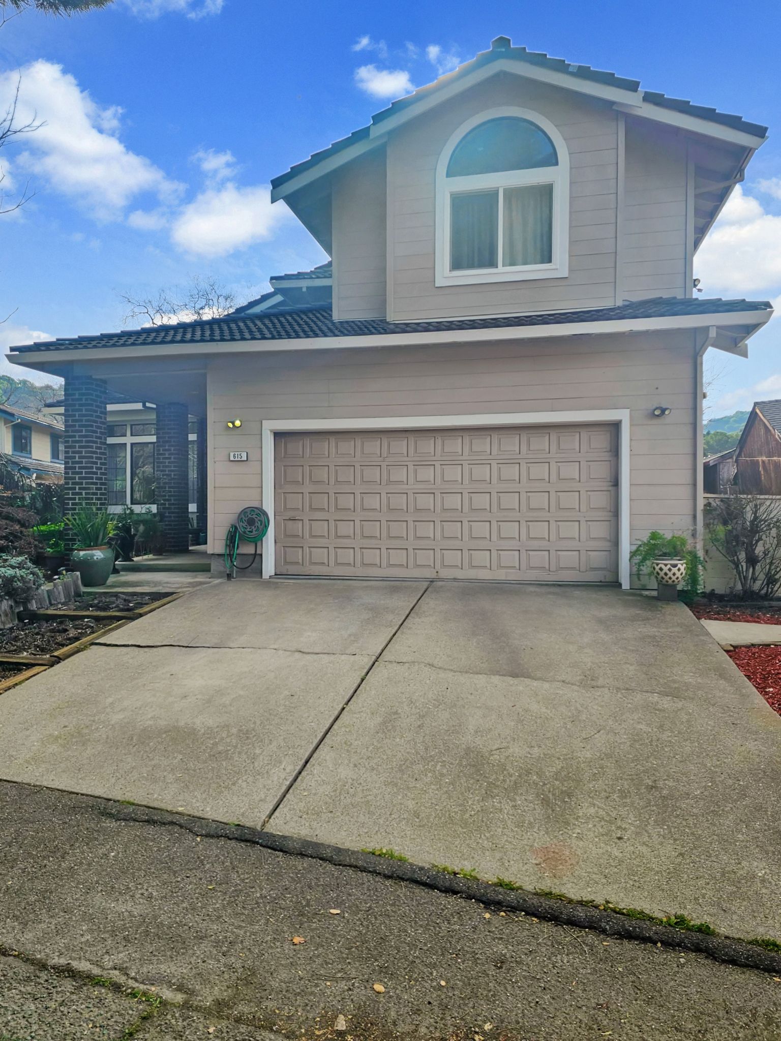 A tan, two-story house with a prominent garage door, concrete driveway, and an arched window on the upper level.