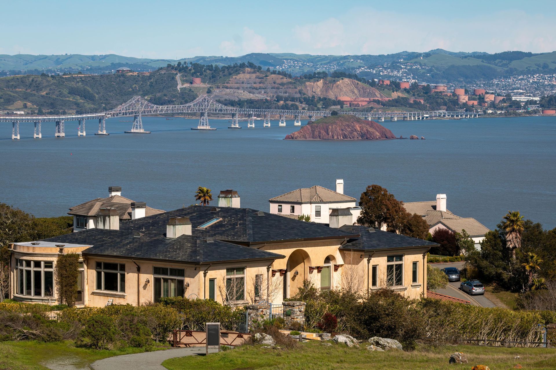 A Mediterranean-style house with a dark roof sits on a grassy hill overlooking the San Francisco Bay and Richmond Bridge.