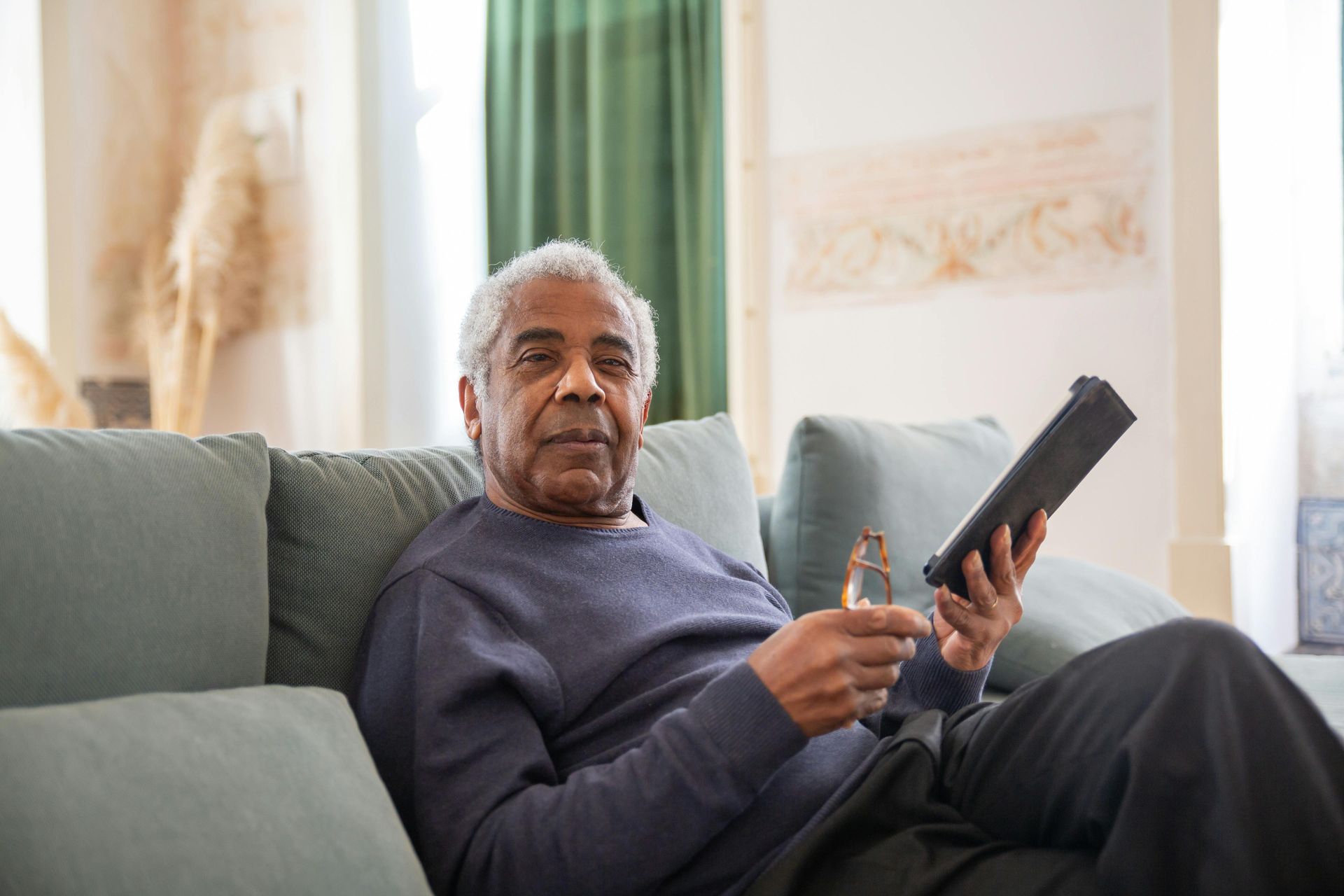 An elderly man is sitting on a couch holding a tablet and a glass of water.