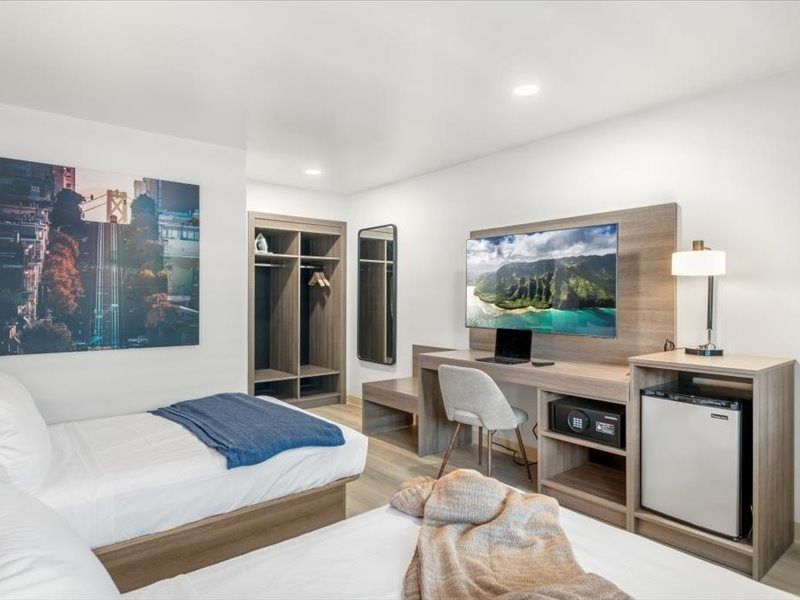 Modern hotel room with bed, desk, chair, TV, open closet, and mountain wall art.