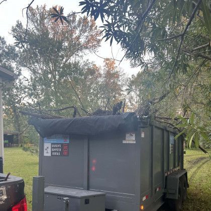 A dumpster trailer is parked in a grassy field with trees in the background.