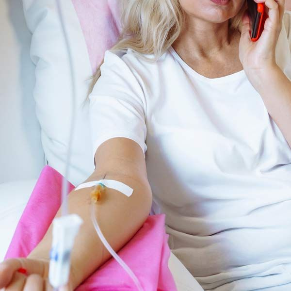 A woman is sitting in a doctor's office with an IV in her arm and talking on a cell phone, representing Advanced IV Therapy for Radiant Wellness