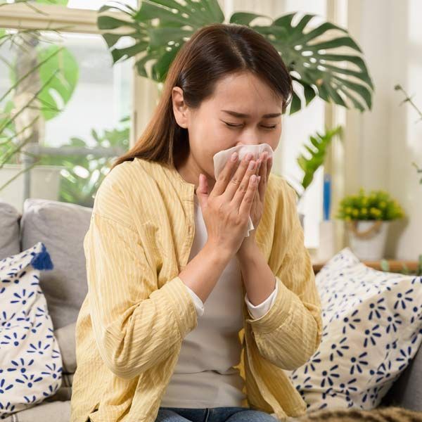 A woman with allergies blows her nose with a napkin while sitting on a couch.