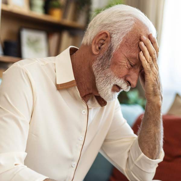 An older man with brain fog and a beard is holding his head with his hand unable to concentrate.