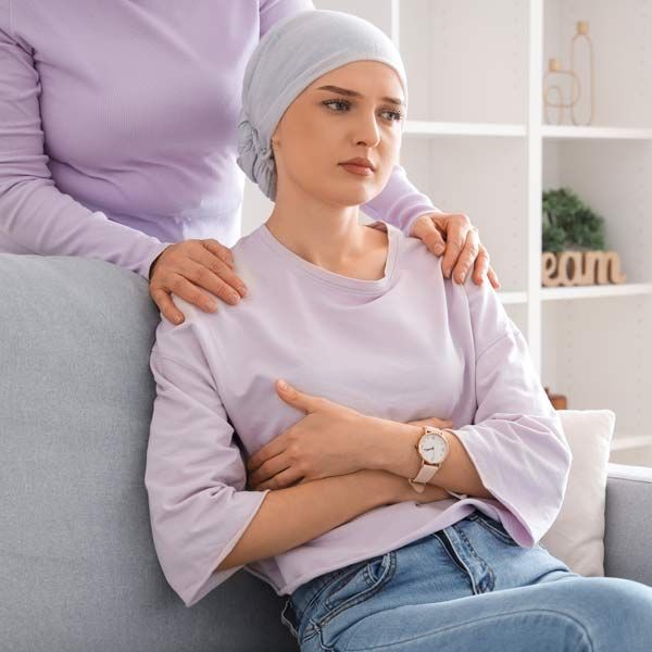 A woman is sitting on a couch with her arm around another woman's shoulder representing Cancer Care