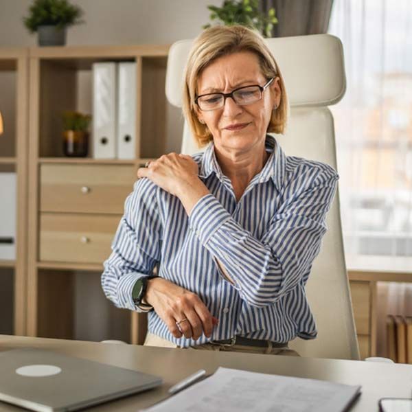 A woman with chronic pain is sitting at a desk, holding her shoulder in pain.