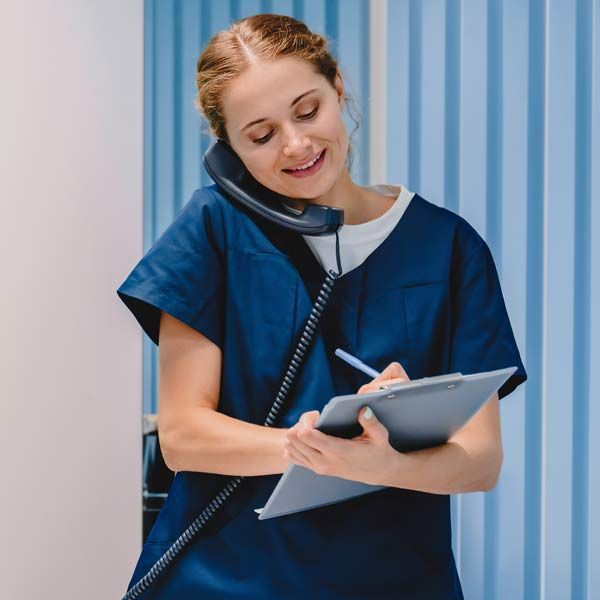 A woman in blue scrubs is talking on a phone and writing on a clipboard. Contact Dr. Kamal Kalsi at Victory Medical Institute for a Consultation