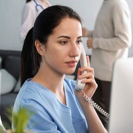 A woman in blue scrubs is talking on a phone in front of a computer, scheduling an appointment for a Victory Medical Institute Consultation