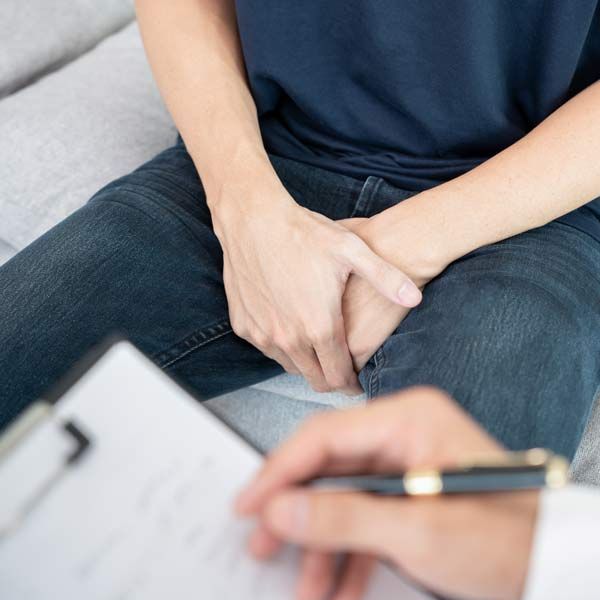 A man is sitting on a couch with his hands on his lap while a doctor writes on a clipboard to treat his Erectile Dysfunction