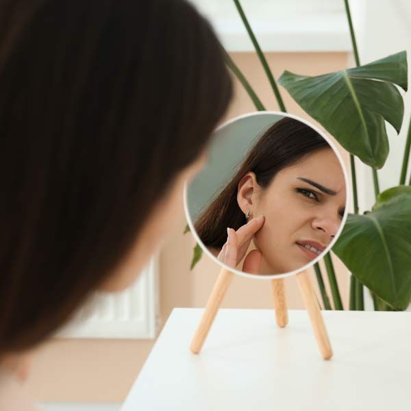 A woman with a hormone imbalance checks a blemish in a tabletop mirror beside a plant.