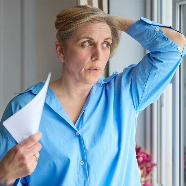 A woman with Menopause in a blue shirt is fanning herself with a piece of paper and looking out a window.