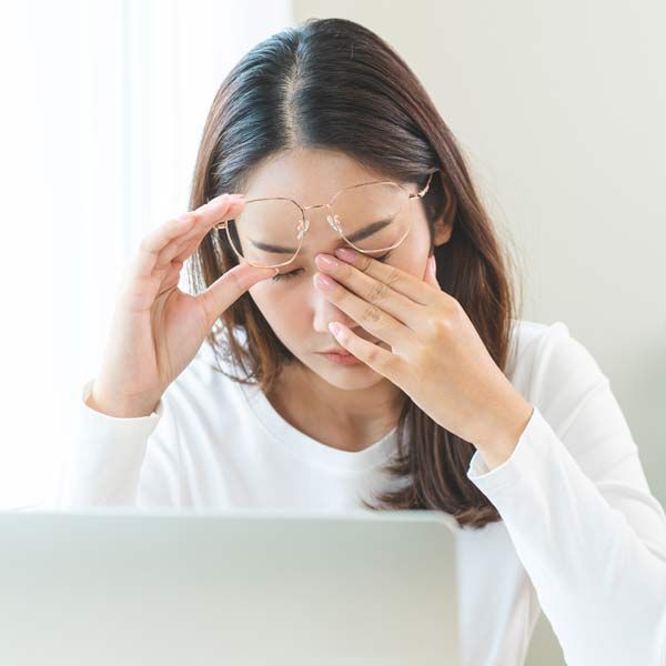 A woman with pain from Migraines wearing glasses is rubbing her eyes while using a laptop computer.