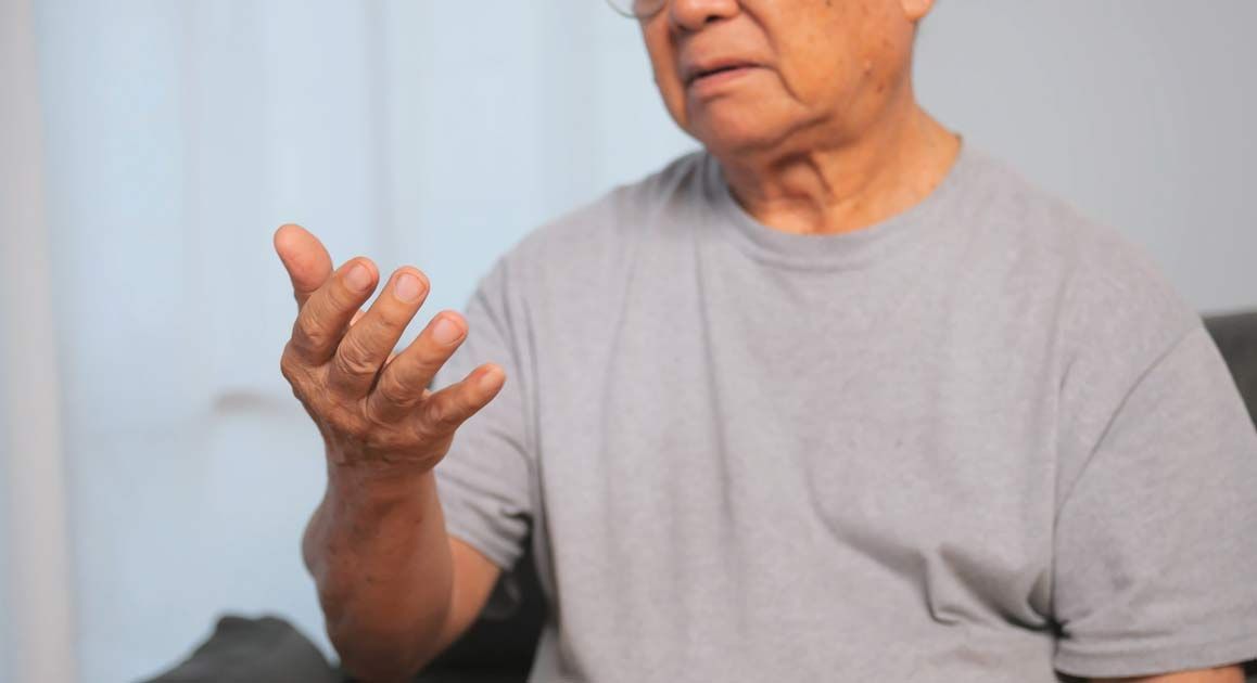 An elderly man is sitting on a couch with trembling hands, needing Neurodegenerative Disease Treatments NJ and NYC - Victory Medical Institute.