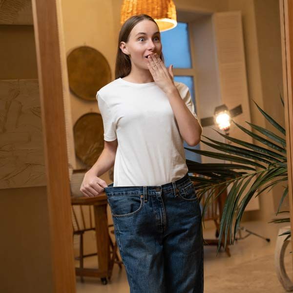 A woman in a white shirt and blue jeans, looking at her weight loss in the mirror from her Peptide Therapy NJ and NYC at Victory Medical Institute