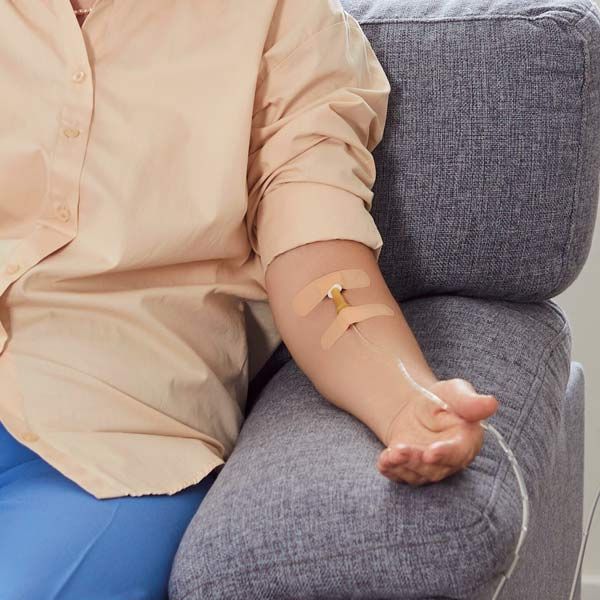 A woman is sitting on a couch with an IV in her arm, representing Quench for Hydration IV Therapy