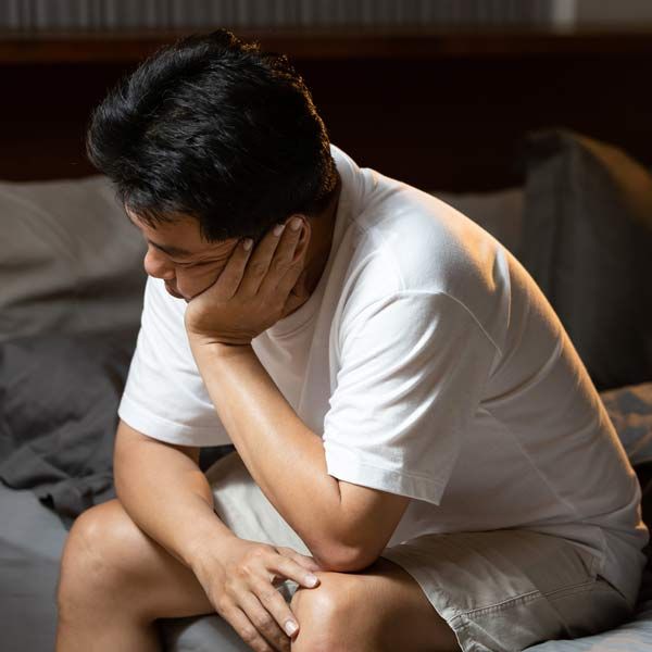 A man in a white shirt is sitting on a couch with his head resting on his hand with Erectile Dysfunction GAINSWave ED Treatments