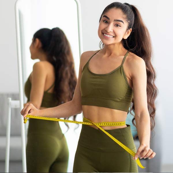 A woman is measuring her waist with a tape measure in front of a mirror, thanks to Advanced Supplements for Weight Loss, Serving Patients in Morris Plains, New Jersey, and Manhattan, New York.