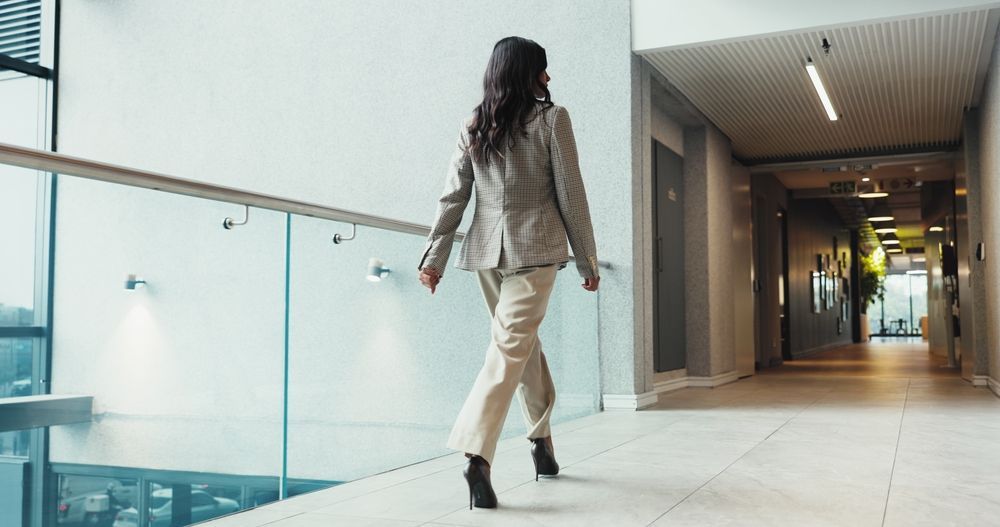 Woman in blazer and pants walks down a bright office hallway, heels clicking.