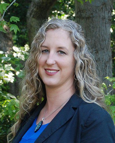 A woman in a blue shirt and black jacket is standing in front of a tree.