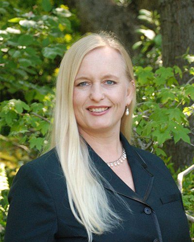 A woman in a black jacket and pearl necklace is smiling in front of a tree.