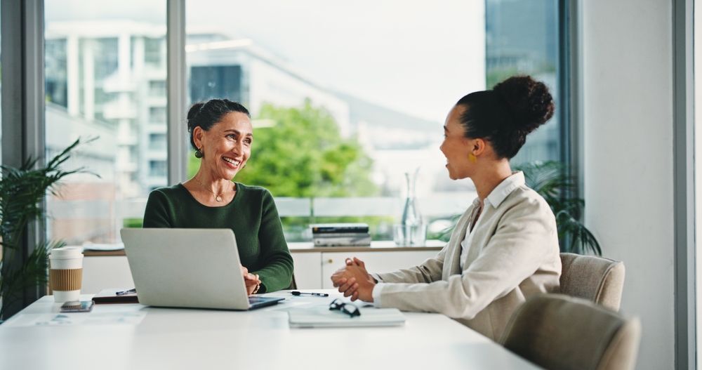 Two professionals in a bright office meeting, smiling and talking while seated at a desk with a laptop.