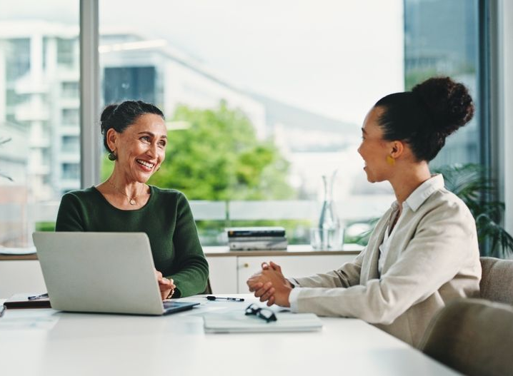 Two professionals have a friendly conversation across a desk in a bright, modern office with a laptop between them.