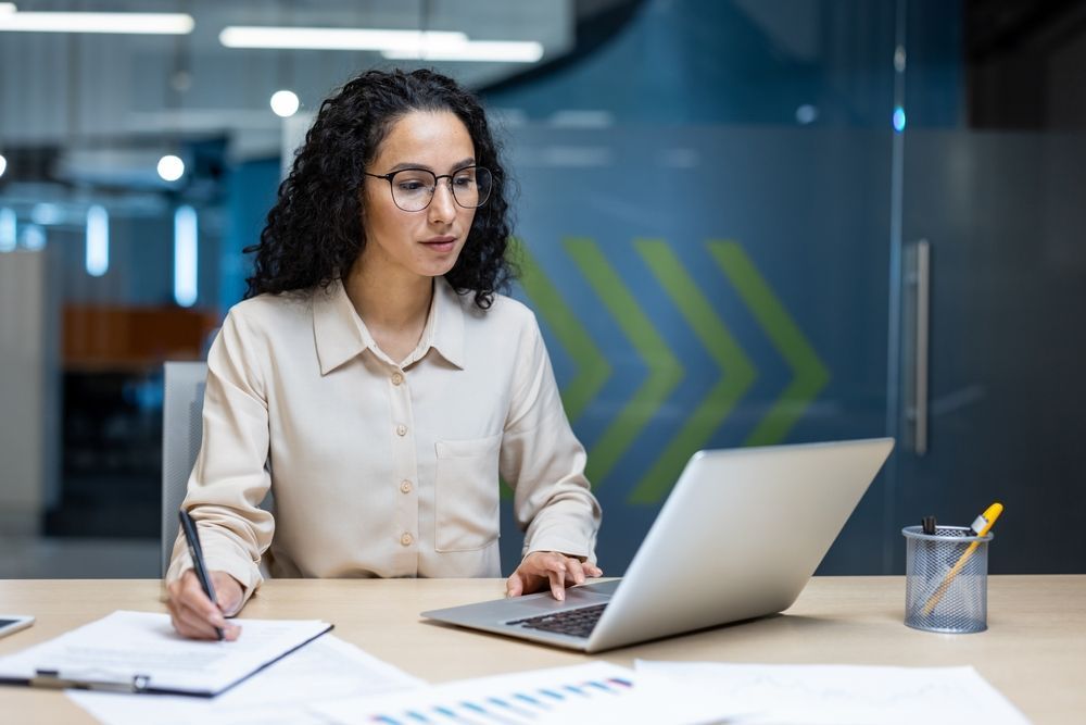 A woman with curly hair and glasses, wearing a cream blouse, works on a laptop and takes notes in an office.