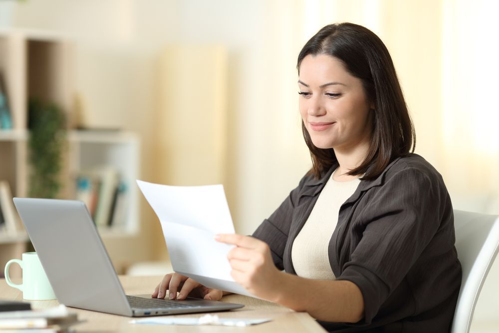 Woman sitting at a desk, looking at a document, laptop open. Sunny room.