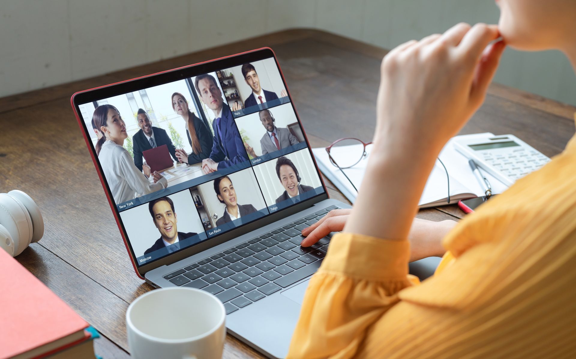 A woman in a yellow blouse, mostly off screen, sits in front of a laptop showing  a virtual meeting with coworkers both working in the office and at home. -Distributed Workforce: How To Create Compelling Benefits