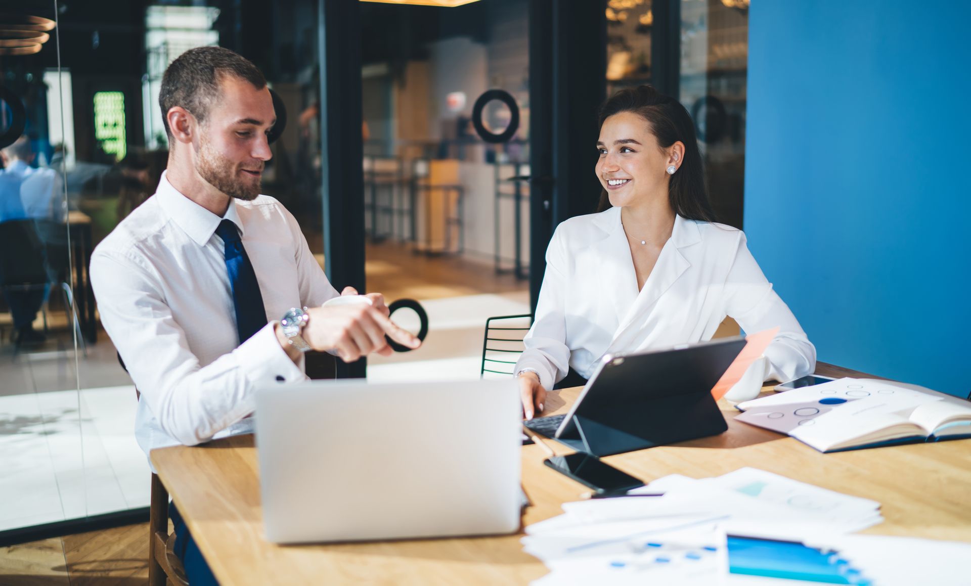 Two colleagues in professional attire collaborate at a desk with a laptop and documents in a modern office space.