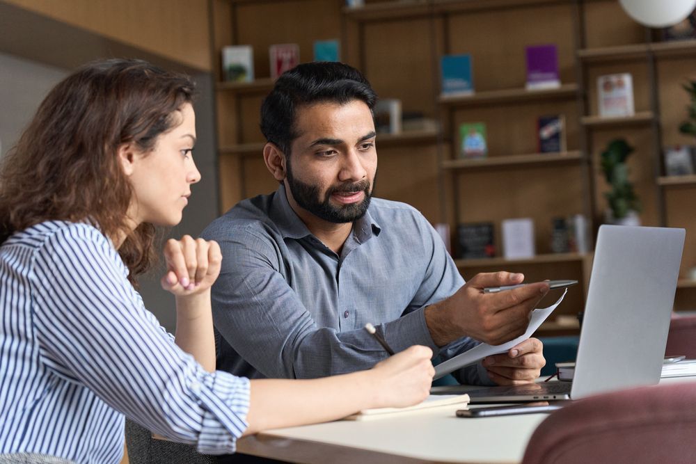 A man and a woman are sitting at a table looking at a laptop.