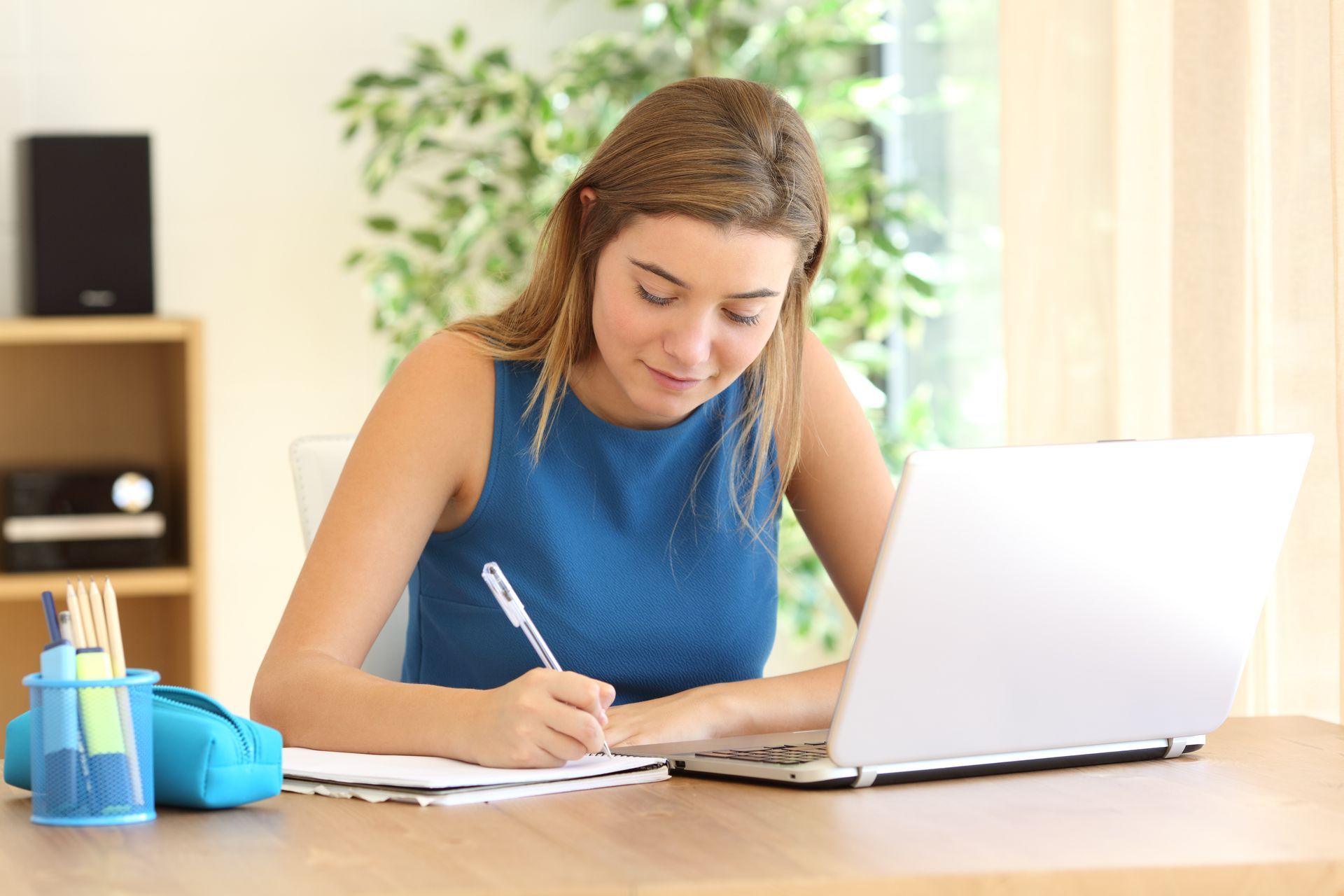 A woman is sitting at a desk with a laptop and writing in a notebook post open enrollment checklist