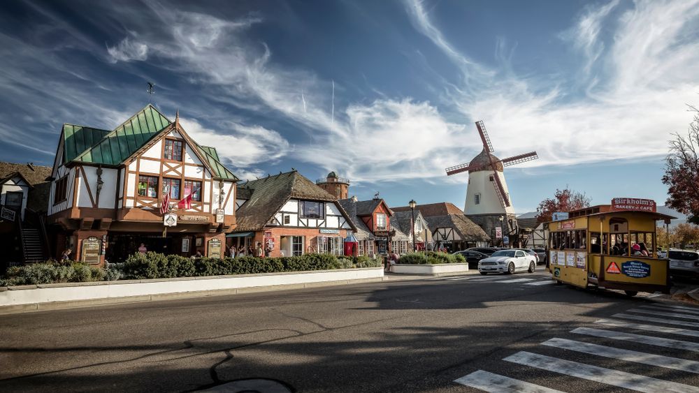 Street scene in Solvang, CA with Tudor buildings, a windmill, blue sky, and a yellow trolley.
