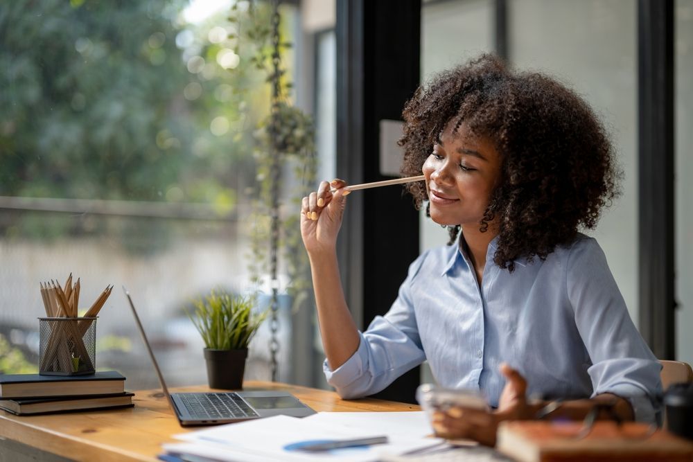 Woman with curly hair, smiling, holding a pencil to her lips, sitting at a desk with a laptop.
