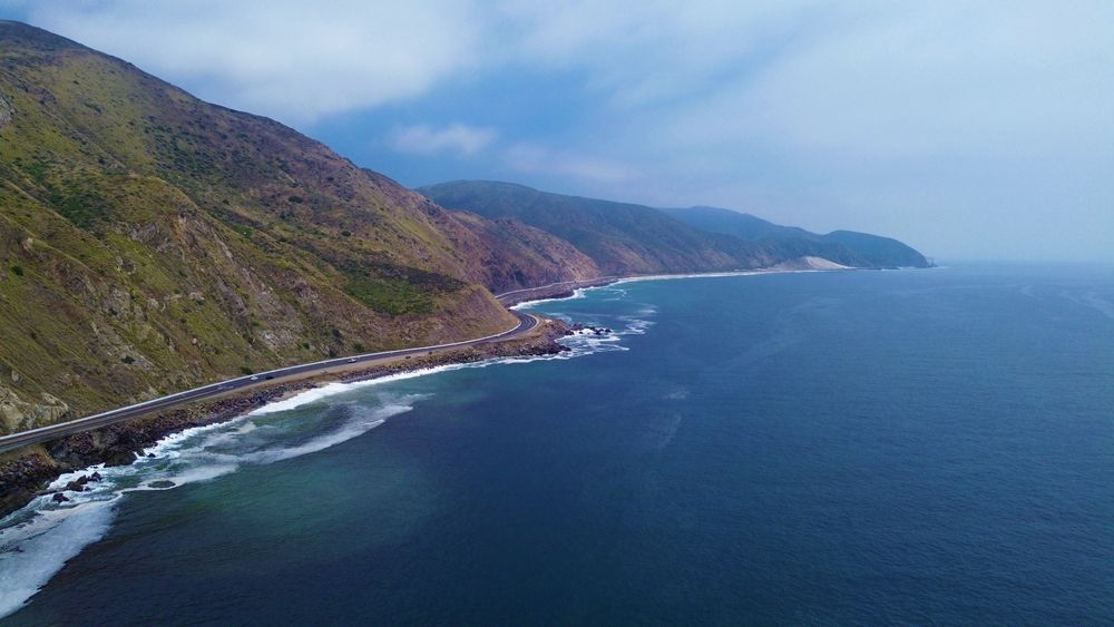 Coastal scene: Mountains meet the sea, road hugs the shore. Blue water, rocky cliffs, cloudy sky.