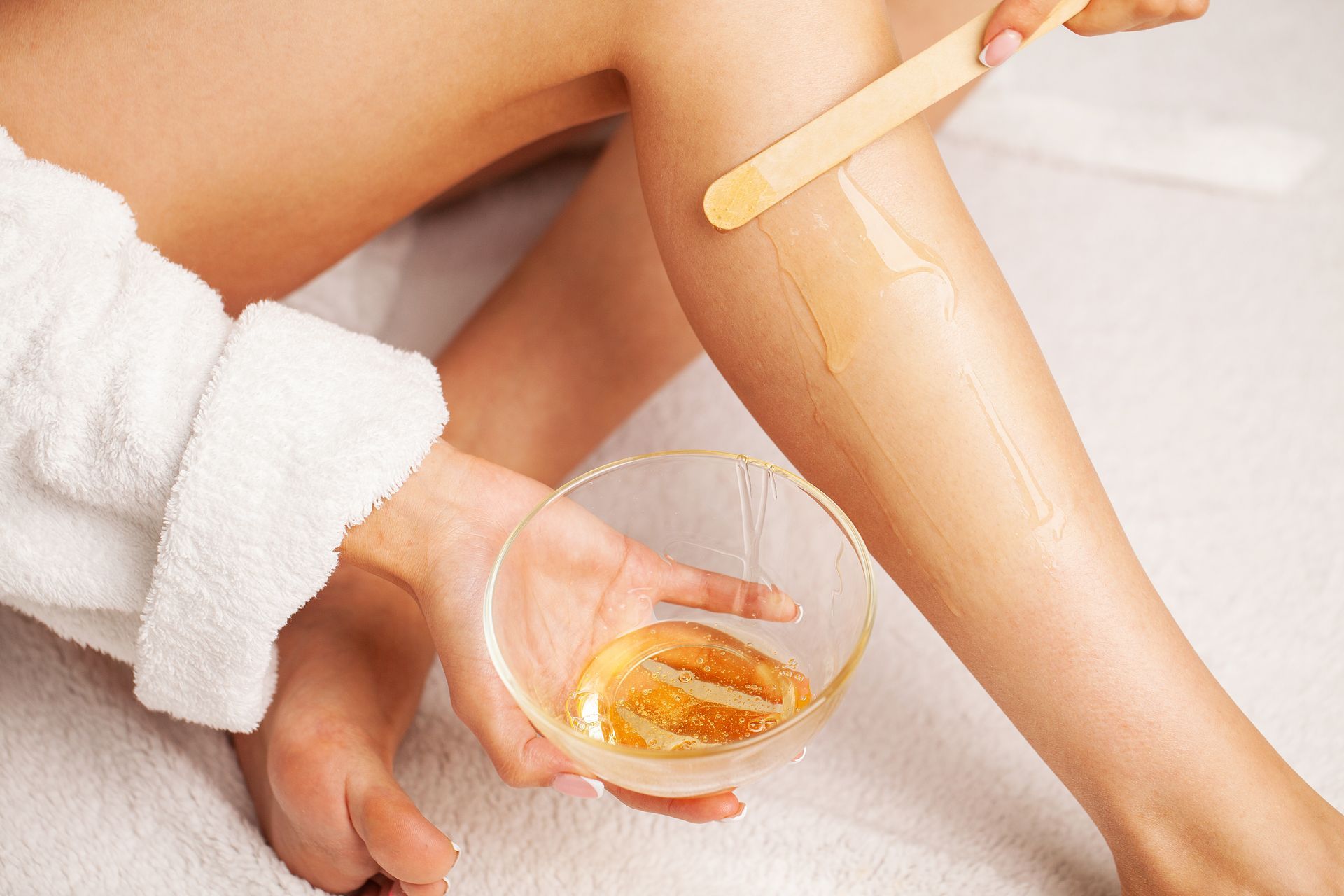A woman is waxing her legs with a wooden stick while holding a bowl of wax.