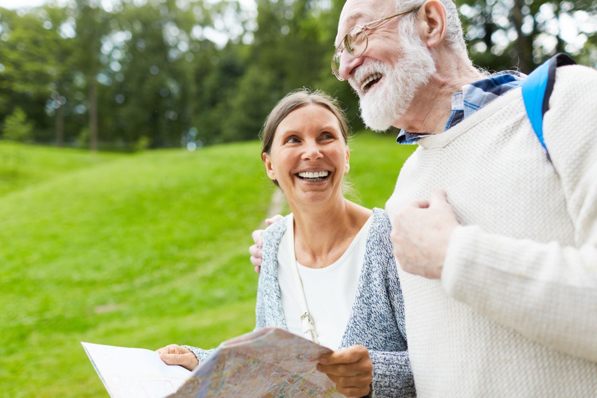 Smiling older couple on a green hill; woman holding map, man with backpack, trees in background.
