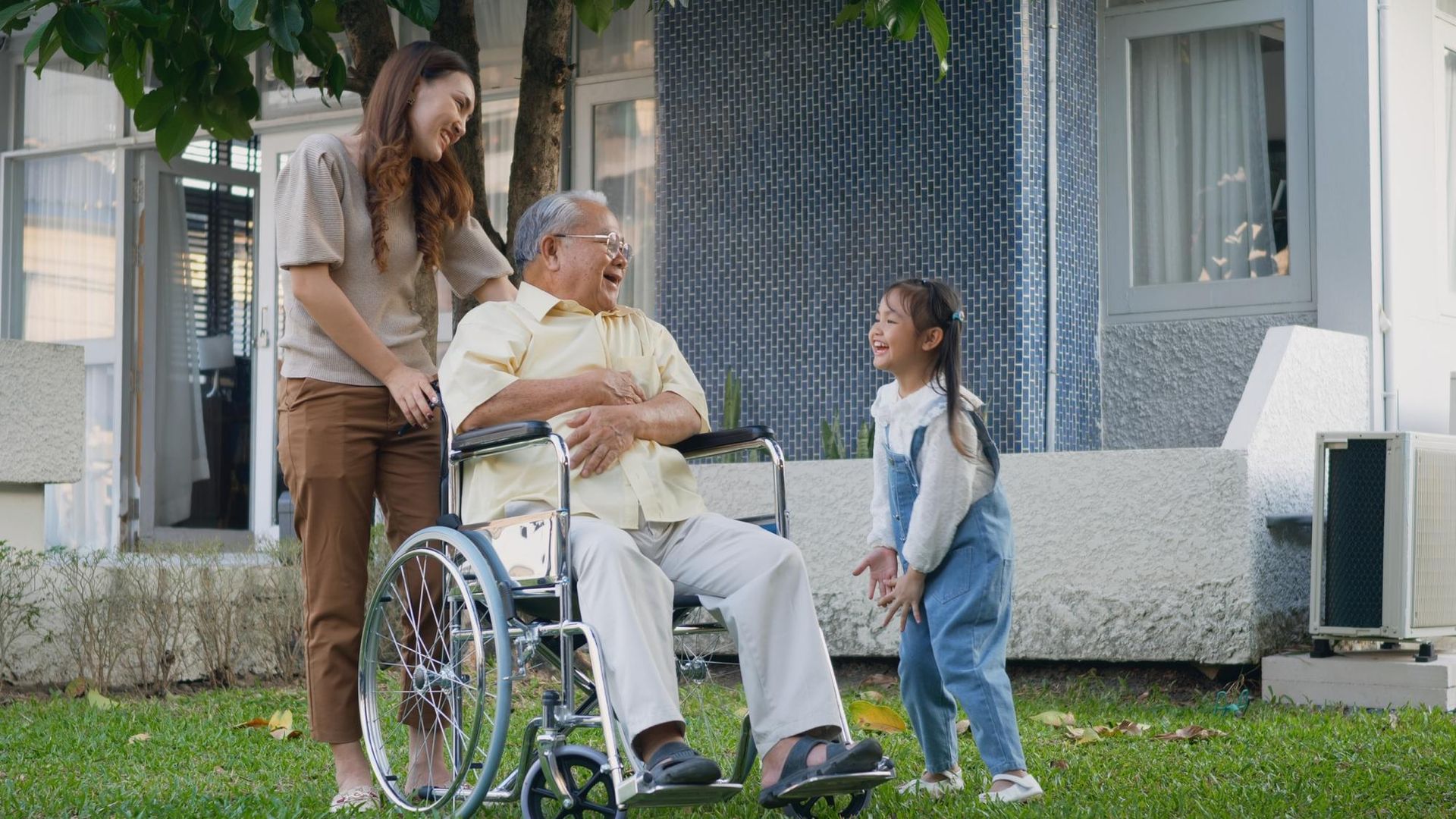 Family, smiling. A grandfather in a wheelchair, a daughter, and granddaughter in a grassy yard.
