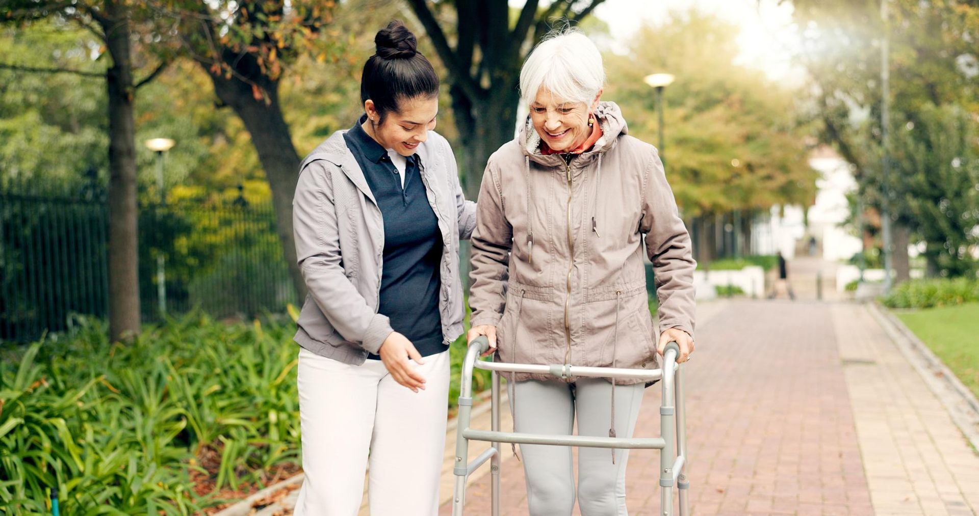 Woman with walker and caregiver walk on path in park. Sun shines, trees in background.