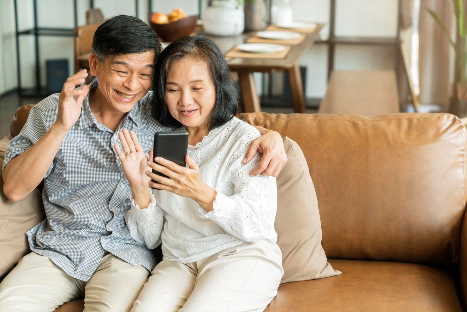 Senior couple using a phone and laptop at a table. Smiling, light-filled room.