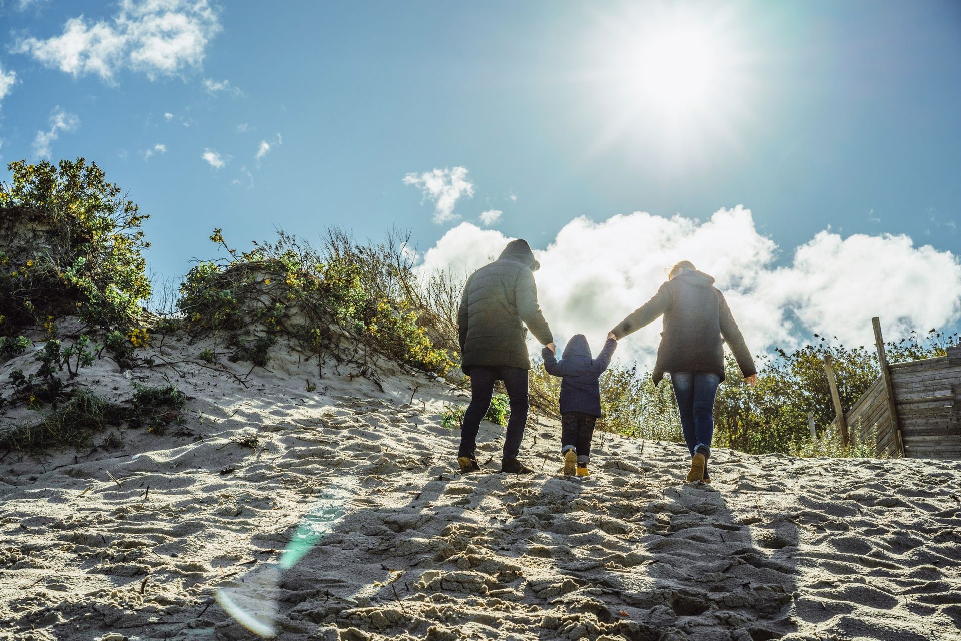 Family walking on a sandy hill, holding hands under a bright sun and blue sky.